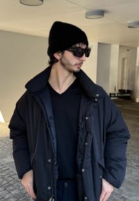 Melnas pūkaina jaka ar augstu apkakli, priekšpusē ar rāvējslēdzēju un spīdīgu apdari; komplektā ar melnu adītu cepuri un saulesbrillēm.