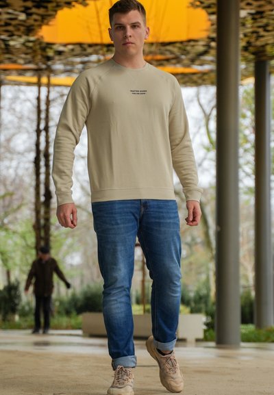 Sudadera beige con cuello redondo, mangas largas y pequeño texto negro en la parte delantera, combinada con jeans azules y zapatillas color beige claro.