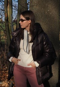 Giacca a vento marrone scuro con cappuccio, maglione crema con accenti grigi e pantaloni rosa chiaro. Materiali strutturati visibili nel tessuto.