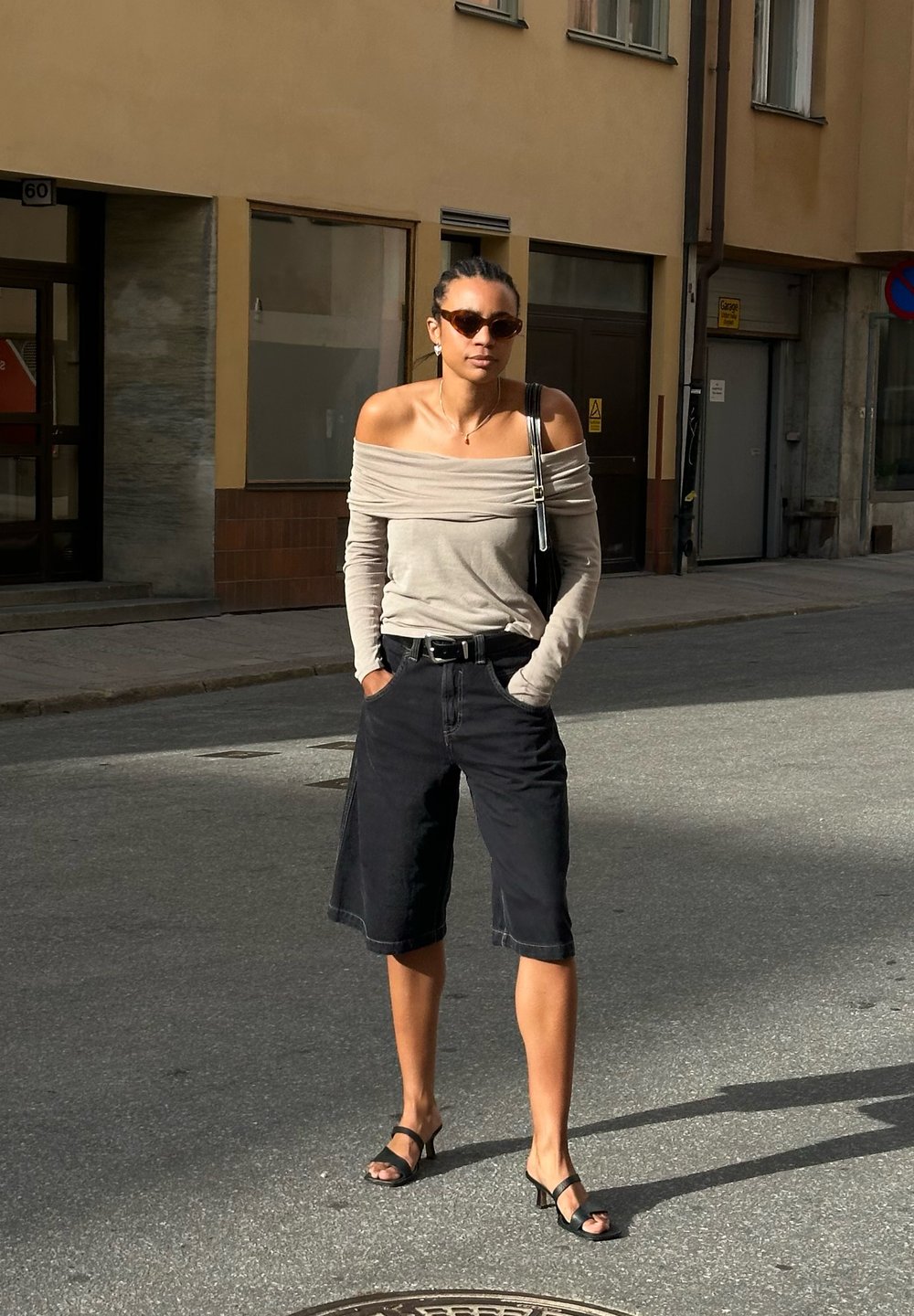 Ελαφρύ γκρι τοπ με ανοιχτούς ώμους, μαύρες τζιν βερμούδες, μαύρες ψηλοτάκουνες σανδάλια και τσάντα χεριού. Λείες υφές και άνετη εφαρμογή.