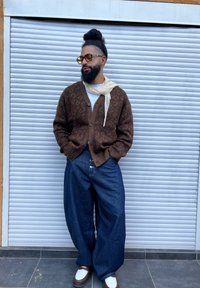 Cardigan cu modele maro peste o cămașă albă, asociat cu pantaloni lejeri din denim albastru și pantofi albi cu maro. Stă în fața unui fundal gri.