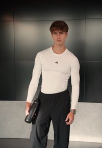 Valge pika varrukaga sportlik särk musta Adidas logoga, sellega on sobitatud mustad lahtised püksid. Modell hoiab käes musta veepudelit.