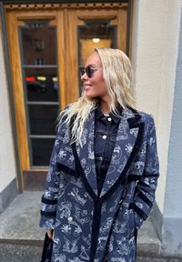 Σκούρο μπλε παλτό με μοτίβα και στυλιζαρισμένα γραφικά, διακοσμημένο με βελούδινη επένδυση και αντίθετες ρίγες. Φορεμένο πάνω από ένα τζιν πουκάμισο, γυαλιά ηλίου.