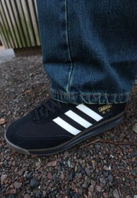 Baskets en daim noir avec trois bandes blanches, logo doré et semelle en caoutchouc foncé. Portées avec un jean bleu, debout sur du gravier.