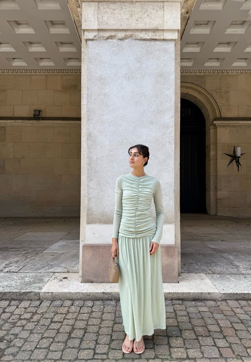 Woman in a pale green ruched top and skirt holding a beige handbag, standing on cobblestones in front of a large stone pillar and archway.