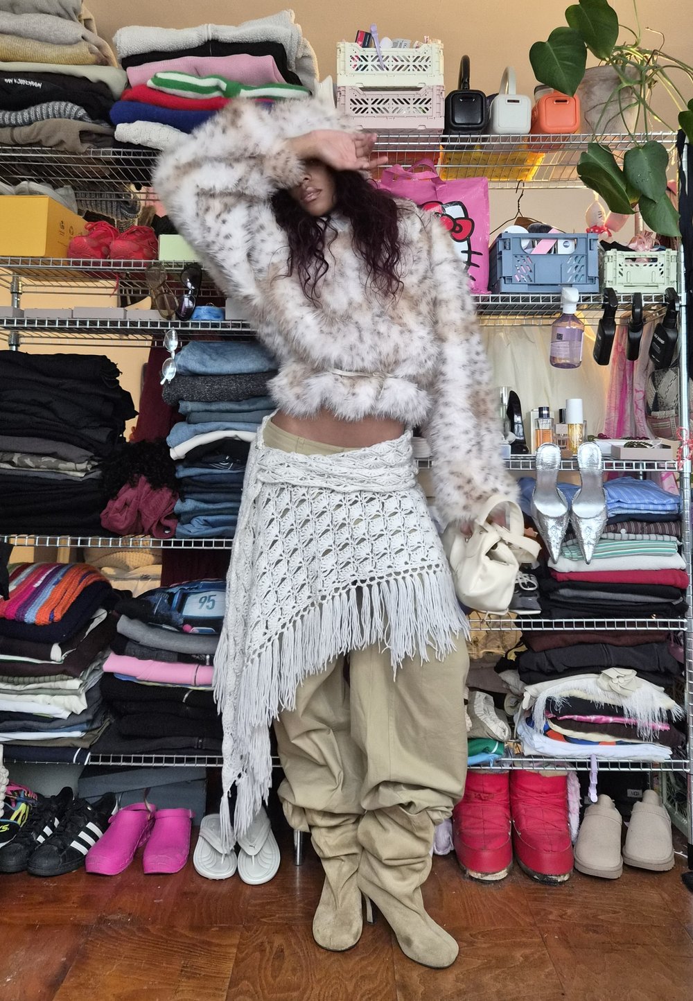 Personne portant un pull en fausse fourrure à motif léopard, un pantalon beige et des bottes, debout devant des étagères métalliques remplies de vêtements pliés et de chaussures.