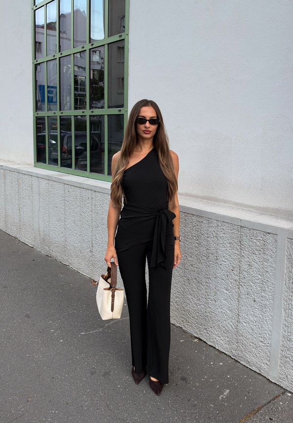 Una mujer con gafas de sol está de pie junto a un edificio, vestida de negro y con un bolso blanco en la mano.