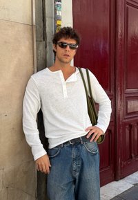 Weißes Henley-Oberteil mit langen Ärmeln und Knopfleiste, lockere blaue Jeans und eine olivgrüne Schultertasche vor einer Backsteinmauer.