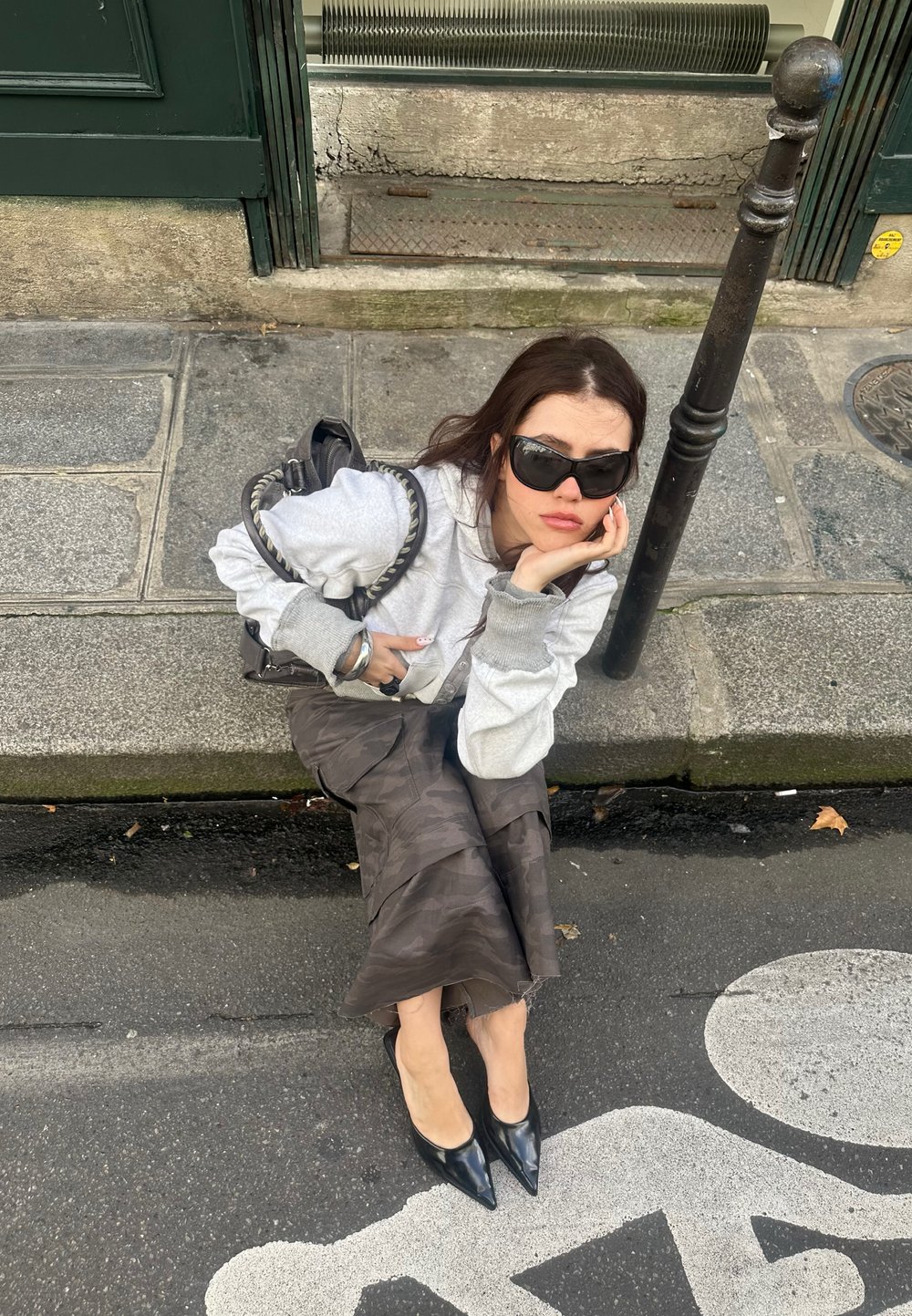 Grijze sweatshirt met geribbelde mouwen, gedragen over een lange rok met camouflagepatroon. Zwarte pumps met spitse neuzen en een gevlochten handtas met een kettinghengsel.