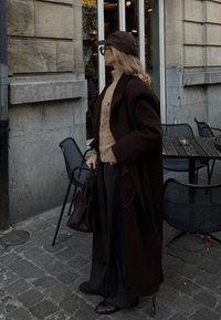 Brun overcoat, strikket beige sweater, mørke bukser og matchende sko. Tilbehør inkluderer en brun kasket og håndtaske. Brostensbelagt underlag og café miljø.