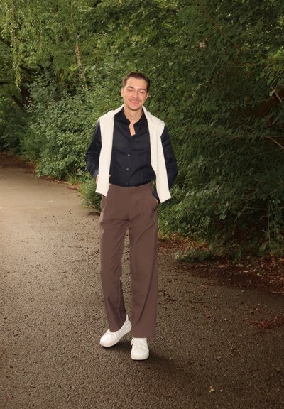 Chaleco blanco sobre una camisa de botones oscura, combinado con pantalones marrones y zapatillas blancas. Rodeado de follaje verde en un sendero.