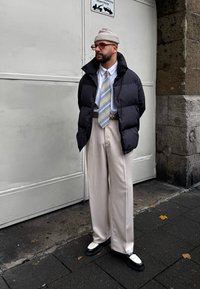 Μαύρο μπουφάν puffer, λευκό πουκάμισο με κουμπιά, μοτίβο γραβάτα, κρεμ παντελόνι φαρδιάς γραμμής και μαύρα παπούτσια με λευκές λεπτομέρειες. Γκρι πλεκτό καπέλο.