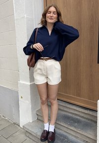 Marinblå tröja med dragkedja i kragen, krämfärgade shorts, brun bälte och en brun väska. Vita ankelstrumpor med mörka loafers, stående på stentrappor.