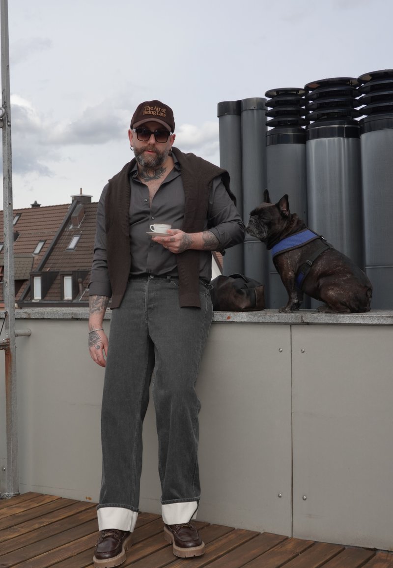 Pán s bradou, slnečnými okuliarmi a šiltovkou drží na streche šálku kávy, vedľa neho sedí čierny francúzsky buldog s modrým postrojom.