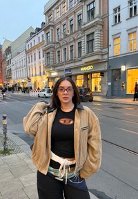 Veston en velours côtelé beige porté sur une chemise noire avec un logo orange, associé à un pantalon foncé et une ceinture à motifs, dans un décor urbain en rue.