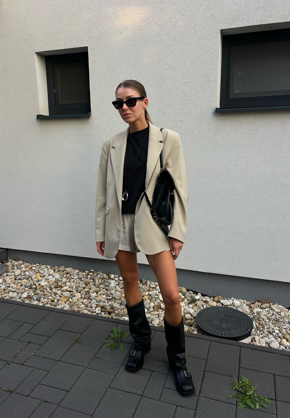 Bež blazer čez črn top, smetana kratke hlače in črne škornje do kolen. Dodatki vključujejo sončna očala in črno torbo. Ozadje iz gramoza.