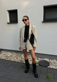 Bež blazer čez črn top, smetana kratke hlače in črne škornje do kolen. Dodatki vključujejo sončna očala in črno torbo. Ozadje iz gramoza.