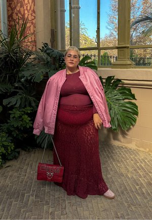 Una mujer lleva una chaqueta bomber de satén rosa sobre una blusa burdeos y una falda de encaje, sosteniendo un bolso rojo con un logotipo metálico.
