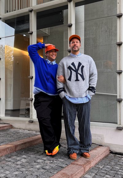 Dos personas de pie sobre adoquines cerca de un edificio; una lleva un suéter azul y gorra naranja, la otra un suéter gris con el logo de NY y pantalones a rayas.