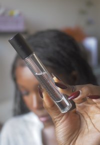 Botella de perfume de vidrio transparente con tapa negra, con una etiqueta con texto en cursiva. Mano sosteniendo la botella con uñas largas de color rojo oscuro.