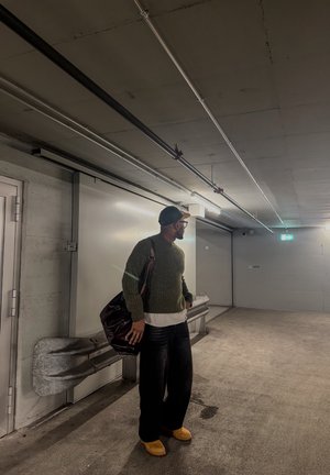 Hombre con suéter verde, pantalones negros y zapatos amarillos, con una bolsa, de pie en un estacionamiento de concreto con poca iluminación.