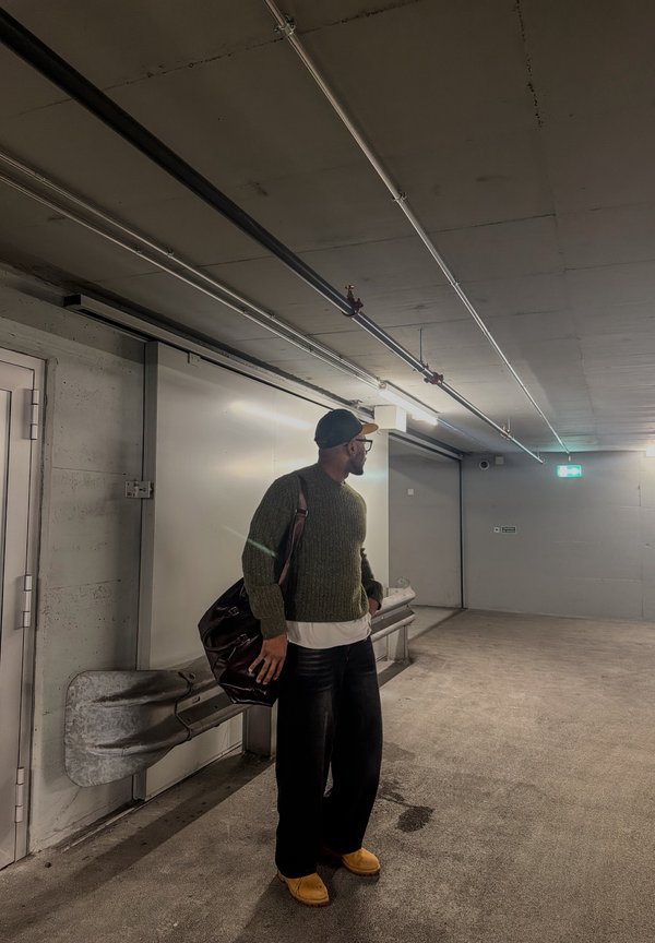 Mann iført grønn genser, svarte bukser og gule sko, bærer en bag, stående i en svakt opplyst betongparkering.