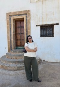 Femme aux cheveux foncés, lunettes, gilet crème, et pantalon vert, debout les bras croisés près des marches et d'une porte en bois décorative.