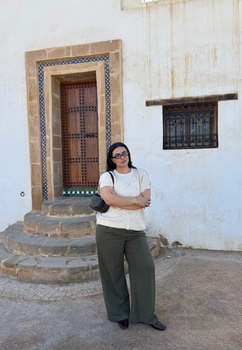 Donna con capelli scuri, occhiali, gilet crema e pantaloni verdi in piedi con le braccia incrociate vicino a gradini e una porta in legno decorativa.