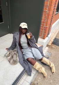 Mujer con trenzas largas que lleva una gorra beige, abrigo gris y botas beige, sentada en la acera, bebiendo café con hielo junto a un bolso peludo.