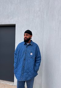 Μπλε καπιτονέ τζιν πουκάμισο με μπροστινή τσέπη και κλείσιμο με κουμπιά, συνδυασμένο με αντίστοιχα μπλε τζιν, φορεμένο με μαύρο καπέλο.