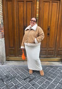 Καφέ σαμωνάκι με λευκό γιακά, ανοιχτό γκρι υφασμάτινη φούστα και πορτοκαλί τσάντα. Στάση σε λιθόστρωτο δρόμο, ξύλινες πόρτες στο φόντο.