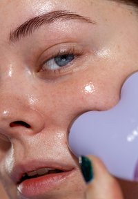 Gezichtreinigingsapparaat in lavendel, met een gestructureerde handgreep tegen glanzende, gehydrateerde huid met zichtbare poriën en lichte reflecties.
