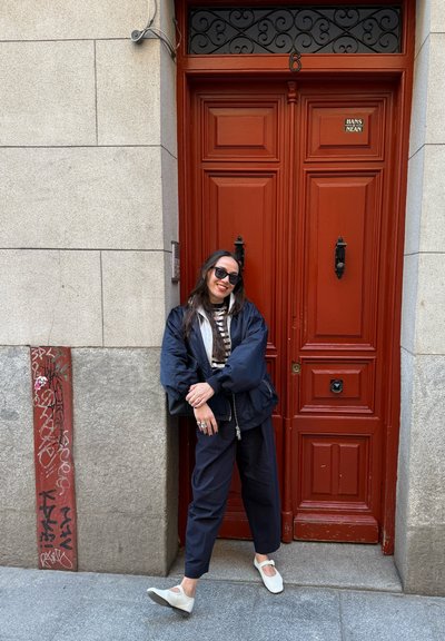 Mujer con chaqueta negra y gafas de sol apoyada en una pared gris junto a una puerta doble roja con manillas negras.