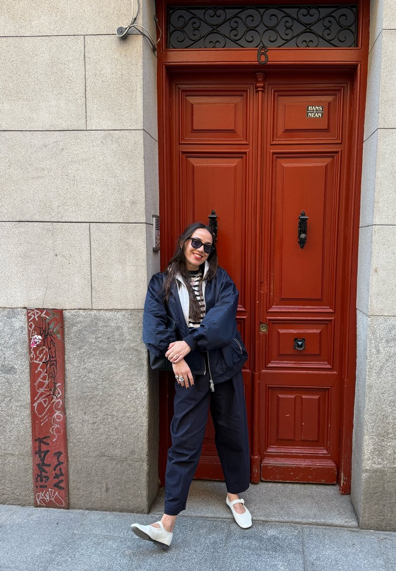 Frau mit schwarzer Jacke und Sonnenbrille lehnt an einer grauen Wand neben einer roten Doppeltür mit schwarzen Griffen.