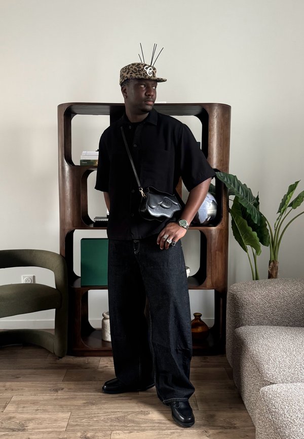 Homme portant une chemise noire et un jean large, casquette à imprimé léopard avec antennes, debout dans un salon près d'une étagère en bois et des plantes.
