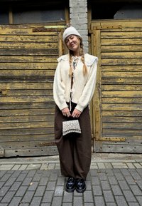Biely pletený cardigan s čipkovým lemom, hnedé široké nohavice, taška s potlačou zvierat, čierne topánky so šnúrkami, vlasy zapletené do vrkočov, stojí proti drevenému pozadiu.