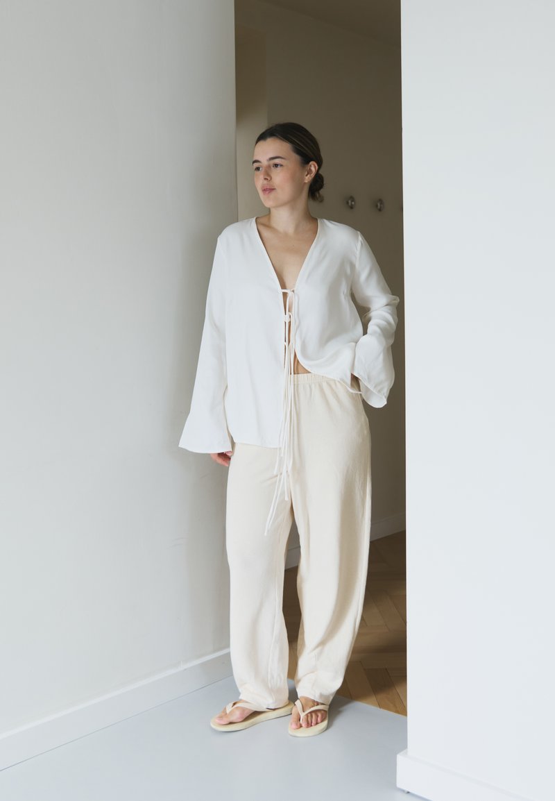 Top branco de manga longa com um decote em V profundo e detalhe de cordão, combinado com calças beiges de corte largo e sandálias simples. Tons neutros.