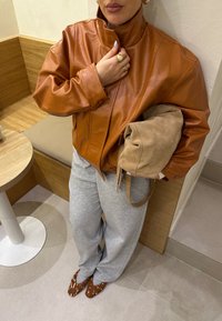 Brązowa skórzana kurtka z wysokim kołnierzem, szare spodnie dresowe i brązowe buty w wzory. Trzyma beżową zamszową torbę. Drewniane tło.