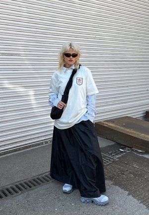 Fehér, oversized póló logóval, kék csíkos ing felett viselve. Fekete hosszú szoknya, fekete keresztpántos táska. szürke sportcipő. Textúrált háttér.