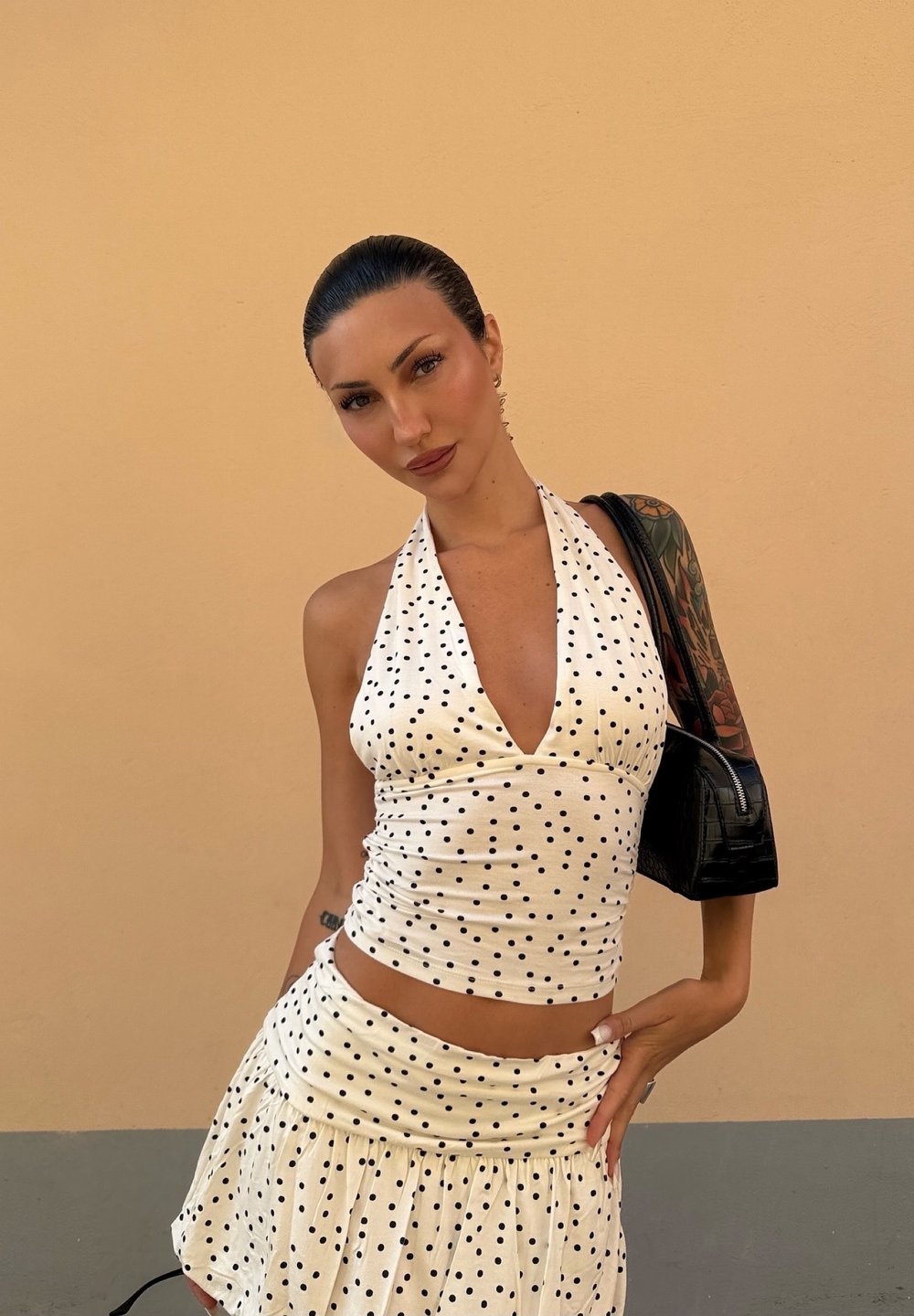 Witte haltertop met zwarte stippen en geplooid ontwerp, gecombineerd met een bijpassende flared rok. Zwarte handtas met een gestructureerde afwerking.