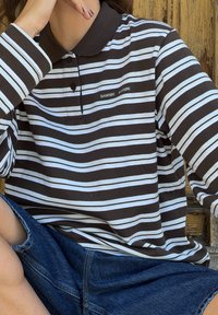 Pessoa vestindo uma camisa polo de manga longa listrada em castanho e branco e calções de ganga azuis, sentada com uma mão repousando perto do rosto.