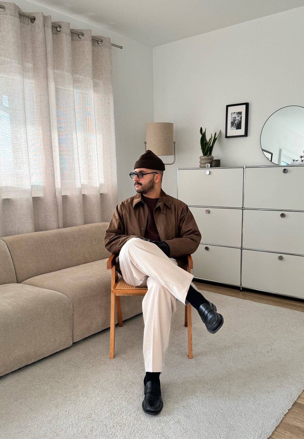 Veste en cuir marron, canapé beige, pantalon blanc et chaussures noires. Rideaux légers, commode minimaliste et plante d'intérieur dans un espace lumineux.