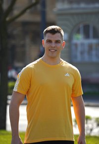 Rumena atletska majica s kratkimi rokavi, ki ima bele tri črtne poudarke na ramenskih delih. Ozadje vključuje zelenje in zgradbe.