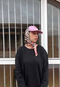 Μαύρο φούτερ, ανοιχτό μπεζ μπλουζάκι από κάτω, ροζ καπέλο με λογότυπο και μαντίλι με σχέδιο ζώων δεμένο στο κεφάλι. Παράθυρο με κάθετους επωμίδες στο βάθος.