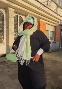 Άτομο ντυμένο με μαύρη στολή, με πράσινο ανοιχτό και λευκό κασκόλ τυλιγμένο γύρω από το κεφάλι, πράσινο καπέλο μπέιζμπολ, κρατώντας πράσινη τσάντα στην πεζοδρόμιο της πόλης.