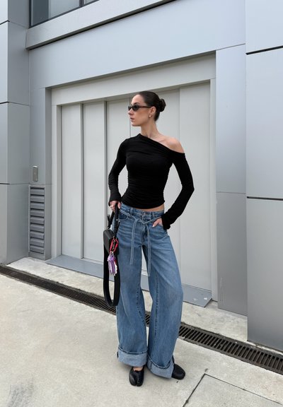 Top negro de manga larga con diseño de un solo hombro, combinado con unos vaqueros de denim azul de pierna ancha. Los accesorios incluyen un bolso de hombro y gafas de sol.