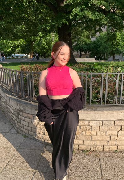 Joven mujer con top corto sin mangas rosa y falda negra sonríe junto a la barandilla de un parque bajo la luz del sol.