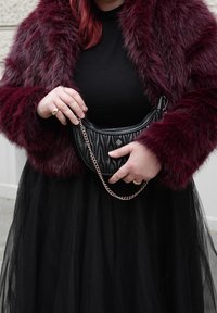 Γυναίκα που φοράει παλτό από συνθετική γούνα σε μπορντό χρώμα και μαύρο φόρεμα κρατά μια μικρή μαύρη τσάντα χειρός με διακοσμητική αλυσίδα σε ασημί χρώμα.