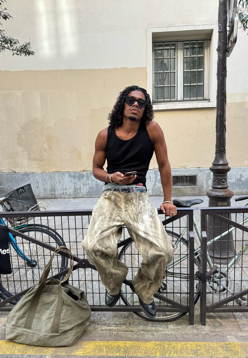 Jeune homme avec des cheveux bouclés et des lunettes de soleil assis sur une clôture urbaine, tenant un téléphone, portant un débardeur noir et un pantalon beige ample à côté de vélos garés.