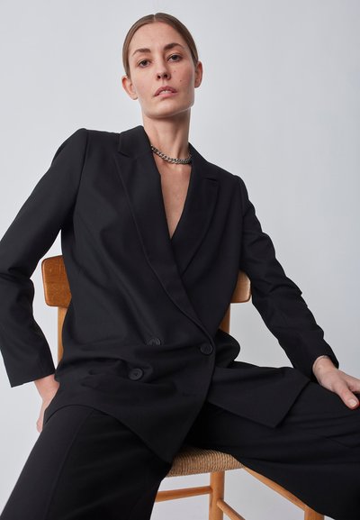 Blazer negro ajustado con un diseño de doble botón, solapas marcadas y textura suave. Combinado con pantalones negros a juego.